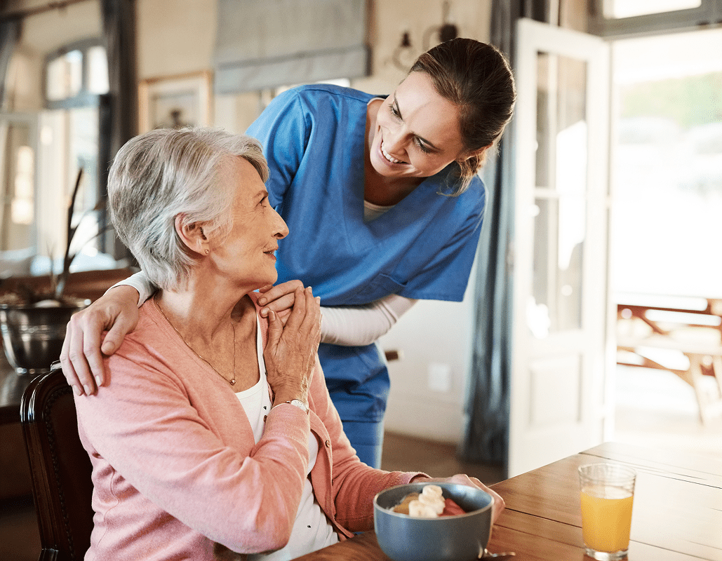 A healthcare worker in blue scrubs gently places her hand on an elderly woman's shoulder and leans in, smiling warmly at her. The elderly woman, seated at a dining table with a bowl of food and a glass of orange juice, looks up at the worker with a caring expression. The setting appears to be a cozy dining area in an assisted living facility with natural light coming through an open door.