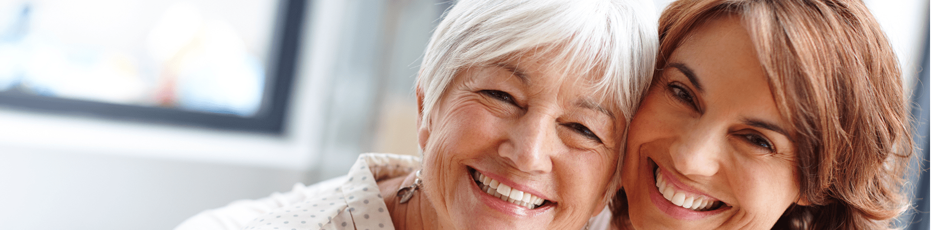Two women, an older woman with short white hair and a younger woman with shoulder-length reddish-brown hair, smiling and leaning close together indoors.