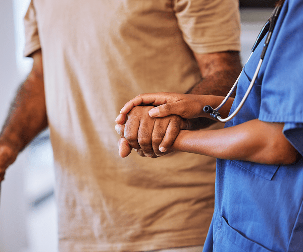 A healthcare worker in blue scrubs gently holds the hand of an older patient wearing a beige shirt, symbolizing care and support at a hospice facility.