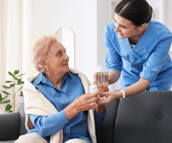 A young female caregiver in blue scrubs offers a glass of water and medication to an elderly woman sitting on a sofa, smiling warmly in a bright, comfortable room.