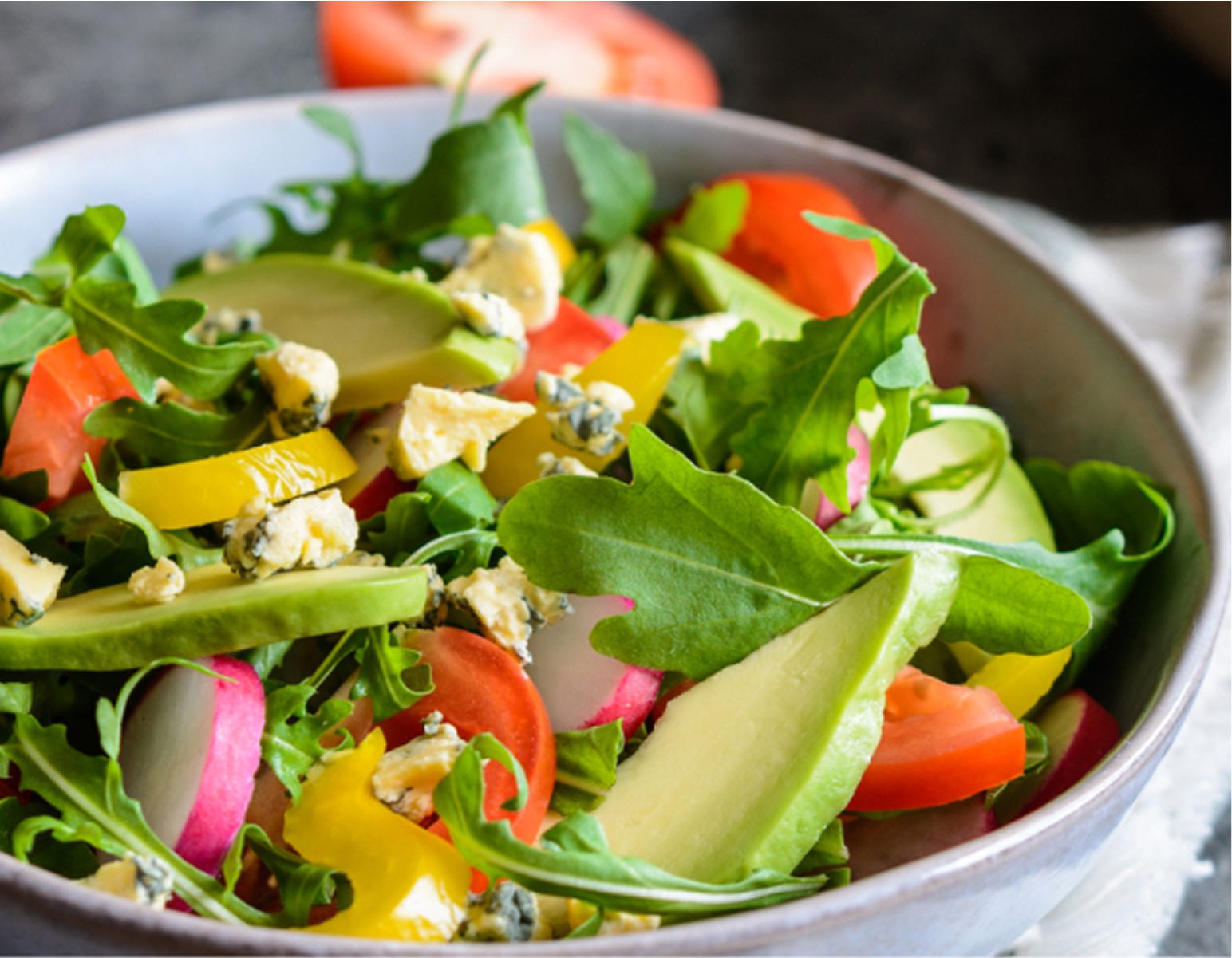 A colorful salad in a bowl with mixed greens, sliced tomatoes, radishes, yellow bell peppers, avocado, blue cheese crumbles, and cucumber slices.