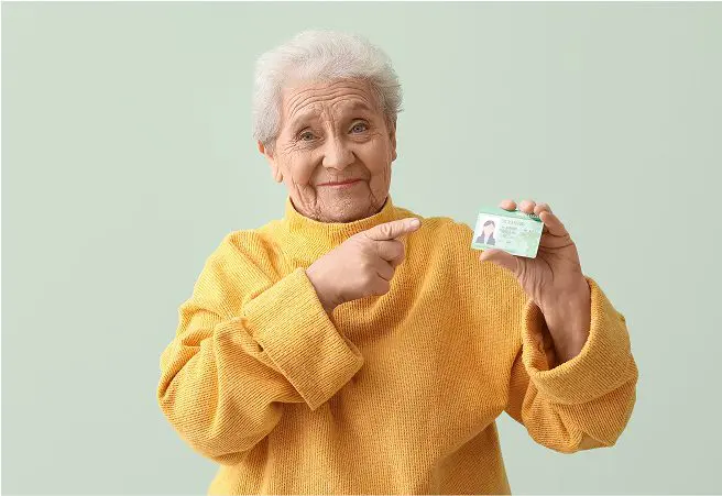 An elderly woman with short white hair, wearing a yellow sweater, points to a driver's license she is holding in her right hand, smiling gently against a light green background.