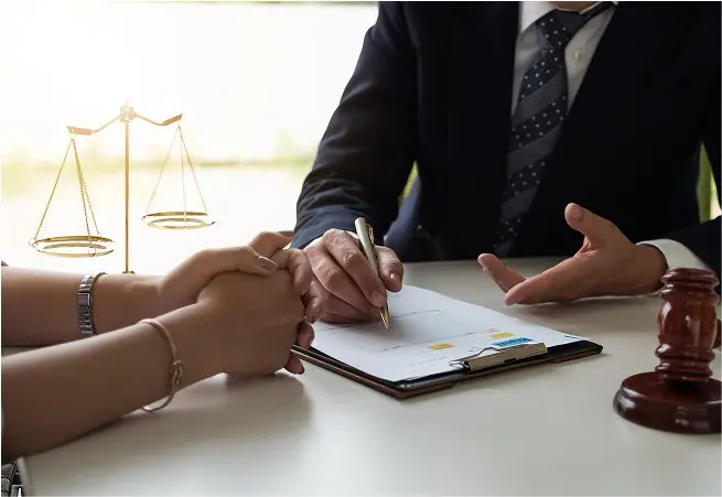 A person in a suit and tie is sitting at a table, signing a document with a gold pen, while another person holds their hands. A gavel and a set of scales are on the table, suggesting a legal or official setting.