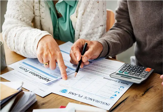 Two people sit at a table reviewing insurance documents; one is pointing at a form while the other holds a pen. A calculator and stacks of paper are also on the table.