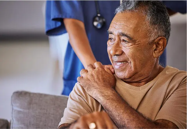 An elderly man smiling and sitting on a couch, with a healthcare worker in blue scrubs standing behind him, gently resting a hand on his shoulder.