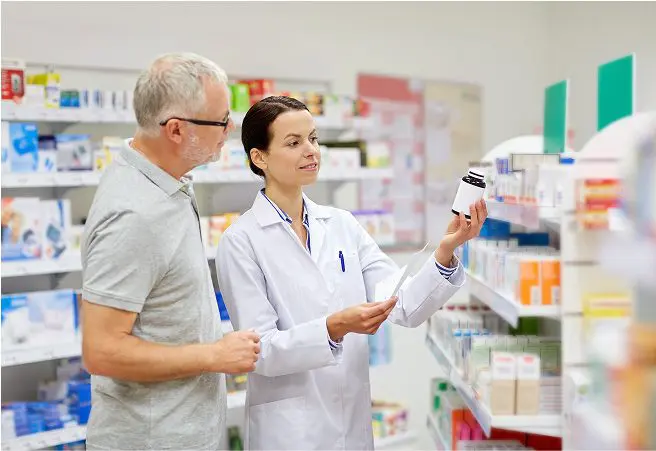 A female pharmacist in a white coat shows a medication bottle to a male customer with gray hair and glasses in a pharmacy aisle filled with various medicine boxes on shelves.