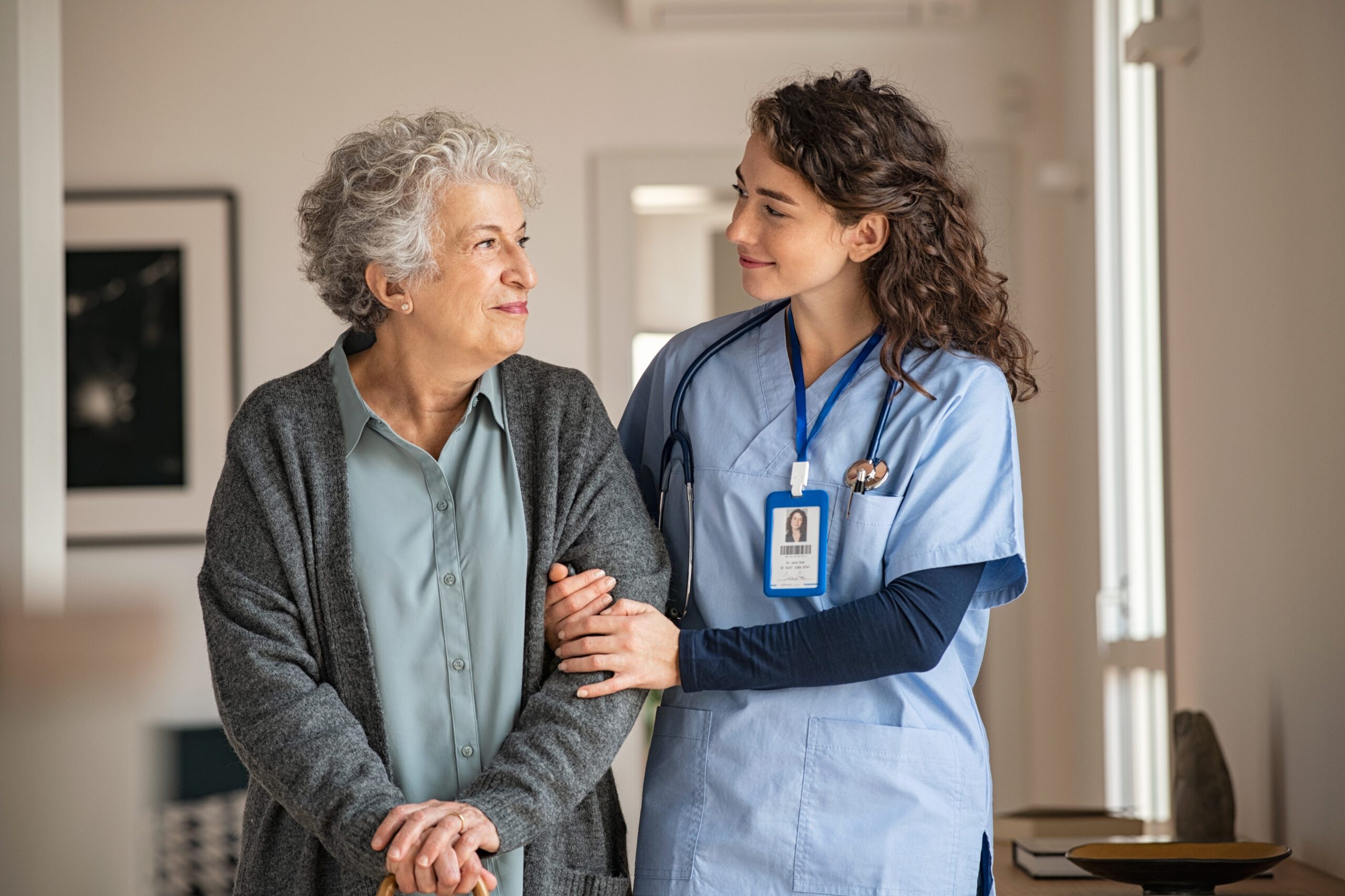 A young female nurse in blue scrubs and a stethoscope around her neck gently holds the arm of an elderly woman with curly gray hair, who is smiling softly. The woman is wearing a gray cardigan over a light blue blouse and is holding a cane. They are indoors in a well-lit room.